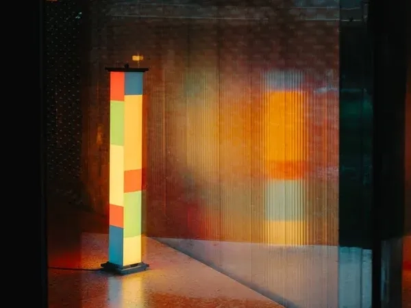 Colorful rectangular floor lamp with red, yellow, blue, and green panels, placed in a modern interior near large windows overlooking urban buildings.