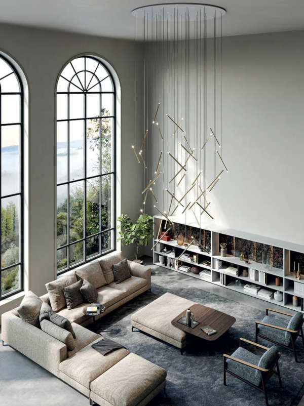 Séjour spacieux avec grande baie vitrée et imposante suspension composée de multiples tiges lumineuses dorées suspendues à différentes hauteurs, éclairant un ensemble de canapés et une table basse sur tapis gris.