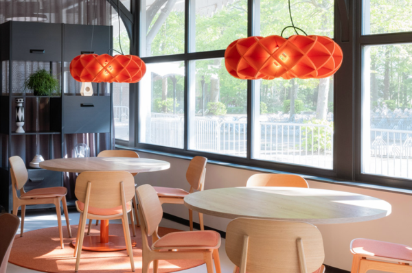 Orange acoustic pendant lights with a sculptural design by Luménear, illuminating a bright room with large windows, light wood round tables, and matching chairs.