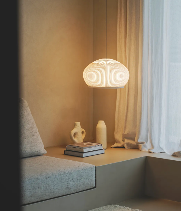 Coin détente avec banquette et livres, éclairé par une suspension textile diffusant une lumière douce près d’un rideau clair.