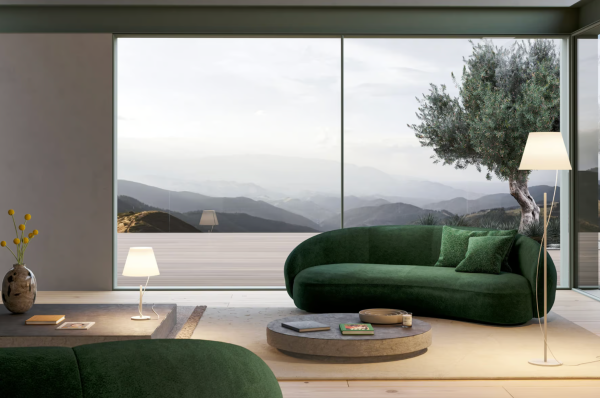 Salon moderne avec canapés verts, éclairé par une lampe de table et un lampadaire blanc, face à une grande baie vitrée donnant sur un paysage de montagne.