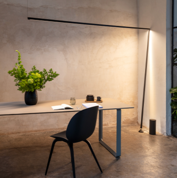 Espace de travail minimaliste avec lampe linéaire noire diffusant une lumière douce vers un mur texturé, table moderne et chaise noire.