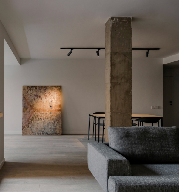 Intérieur moderne avec canapé gris et colonne en béton, éclairé par un rail de spots au plafond et une lumière dirigée mettant en valeur une toile posée au sol.