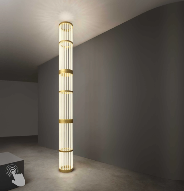 Colonne lumineuse verticale en métal doré et diffuseurs tubulaires, éclairant du sol au plafond dans un espace intérieur minimaliste.