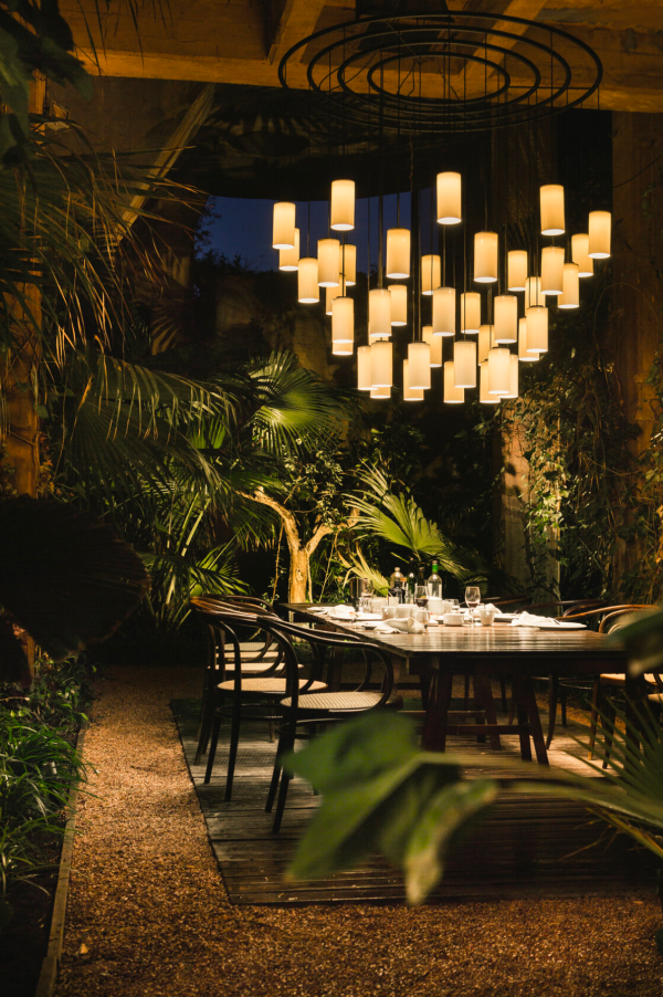 Grand lustre contemporain aux multiples cylindres lumineux éclairant une table à manger en extérieur, créant une ambiance chaleureuse au milieu de la végétation.