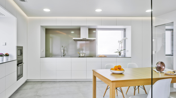 Éclairage encastré et bandeau lumineux au plafond illuminant une cuisine blanche minimaliste avec table en bois.