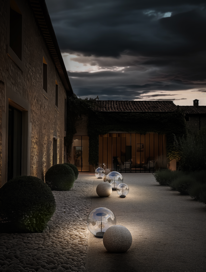Allée extérieure d’une maison en pierre éclairée au crépuscule par des luminaires sphériques en verre de la marque Arkoslight, créant une ambiance élégante et chaleureuse.