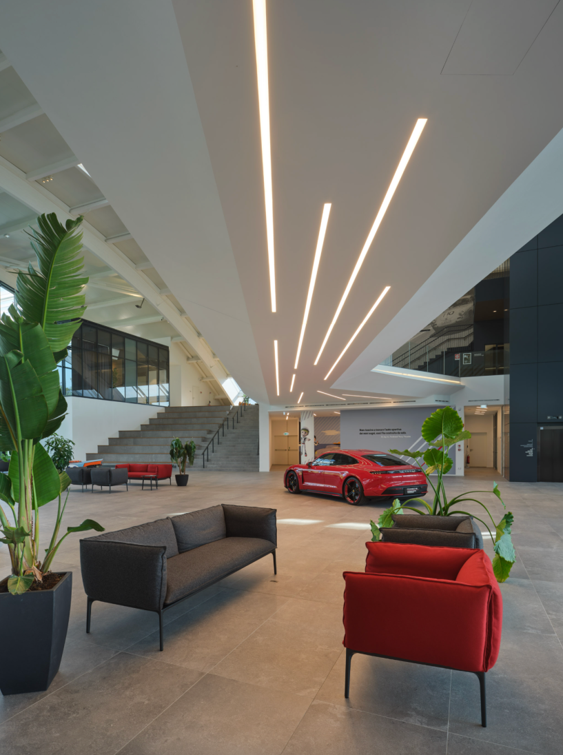 Luminaires linéaires Kalis de Intra Lighting intégrés au plafond d’un vaste hall moderne, éclairant un espace avec mobilier contemporain, grandes plantes et voiture de sport rouge exposée.