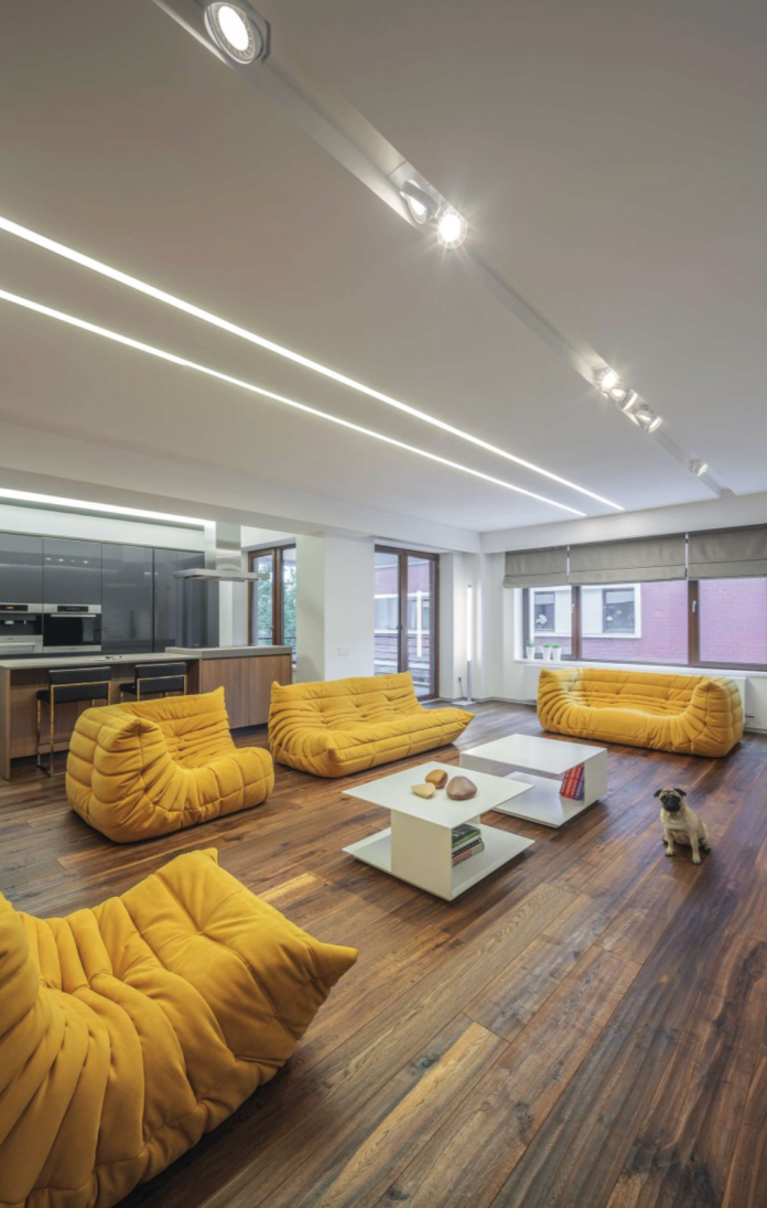 Luminaires linéaires Kalis de Intra Lighting intégrés au plafond d’un salon contemporain avec parquet en bois, canapés jaunes design et table basse moderne.