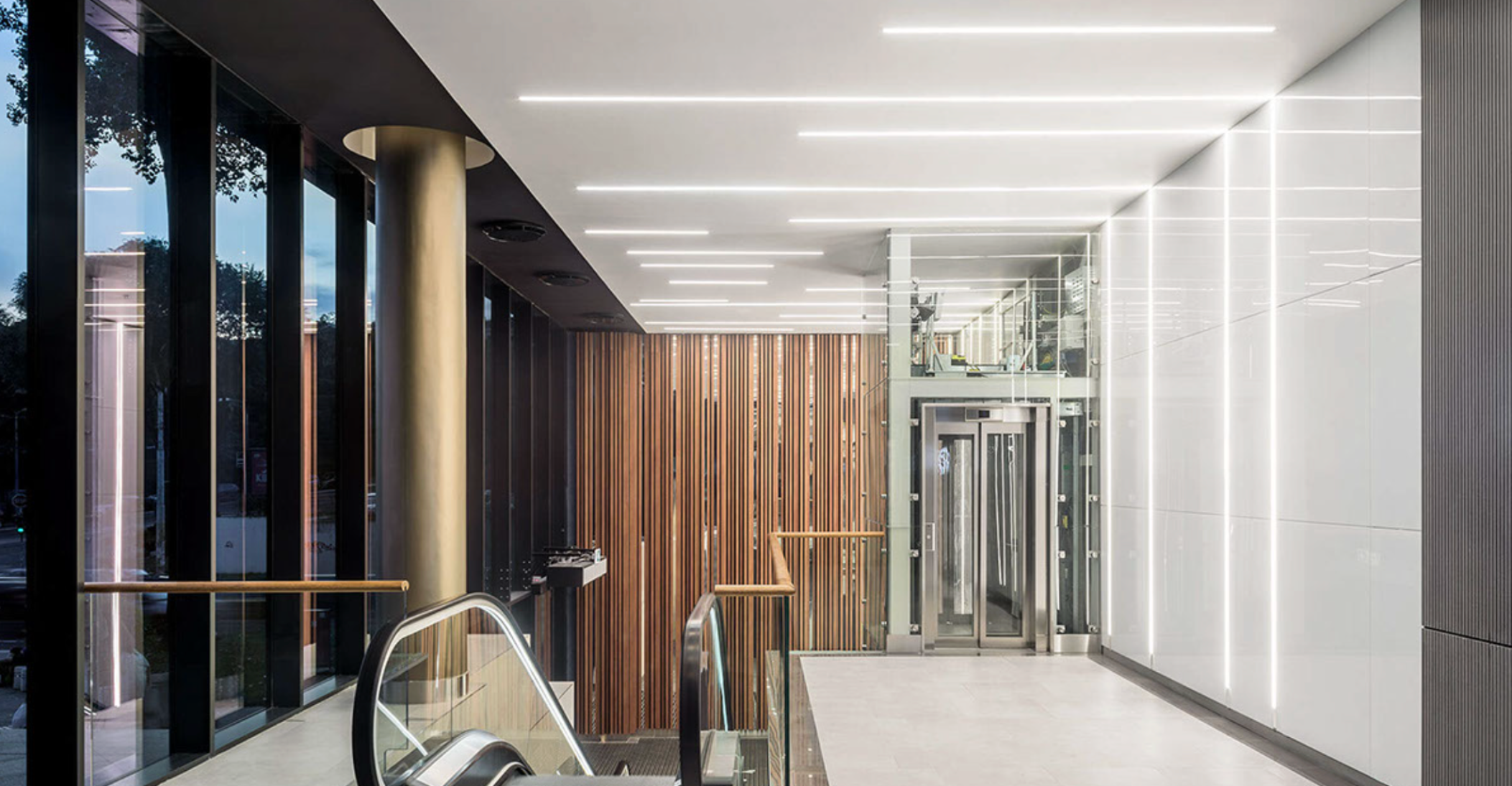 Luminaires linéaires Kalis de Intra Lighting intégrés au plafond d’un hall contemporain avec ascenseurs, murs en bois et assises modernes, créant une ambiance élégante et lumineuse.