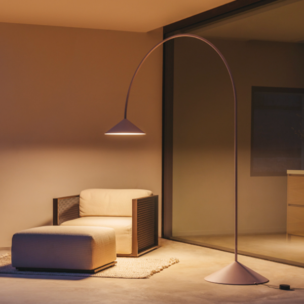 Lampadaire au design courbé avec abat-jour conique diffusant une lumière chaude, placé près d’un fauteuil et d’un pouf dans un intérieur moderne.