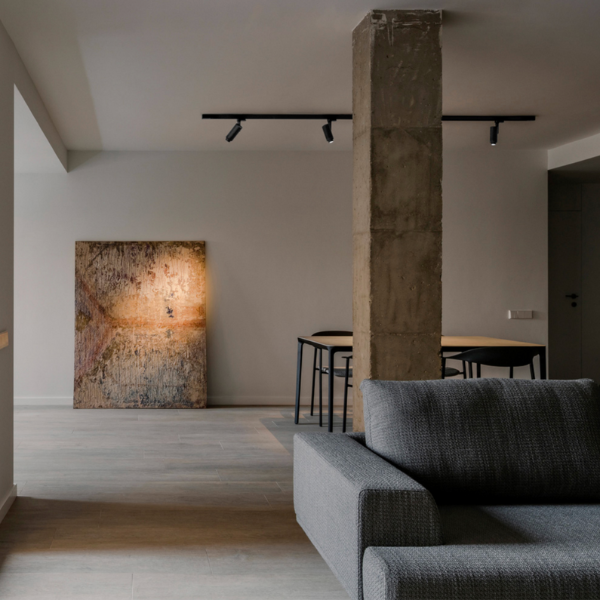 Intérieur moderne avec projecteurs sur rail au plafond éclairant une œuvre d’art, colonne en béton brut et canapé gris.