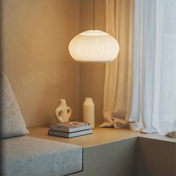 Suspension lumineuse au design textile diffusant une lumière douce, installée dans un coin lecture minimaliste avec coussin, rideaux clairs et objets décoratifs en céramique.
