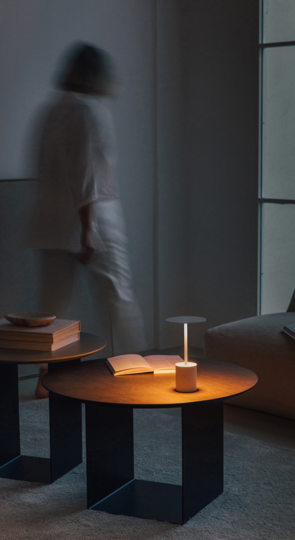 Minimalist portable table lamp with a cylindrical base and horizontal light disc, casting a warm glow on a wooden coffee table in a softly lit setting.