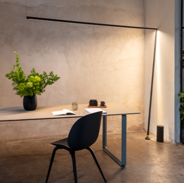 Minimalist floor lamp consisting of a horizontal light rod attached to a vertical black pole, casting indirect light onto a textured wall, placed beside a contemporary desk.