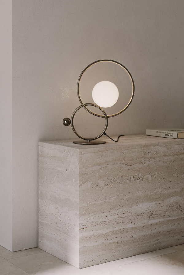Sculptural table lamp composed of two interlocking metal rings, featuring a white opaline glass globe light, set on a round base in a minimalist light-stone interior.