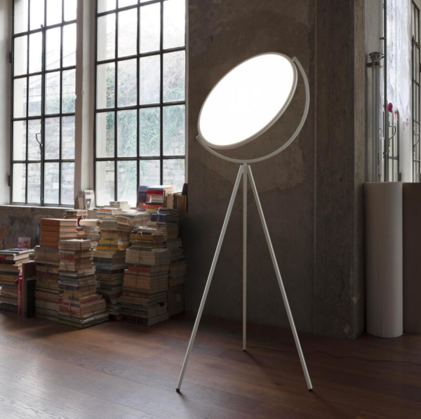 Designer floor lamp on a white tripod base, featuring a large adjustable luminous disc providing even light, set in an industrial-style interior with stacks of books and a large window.