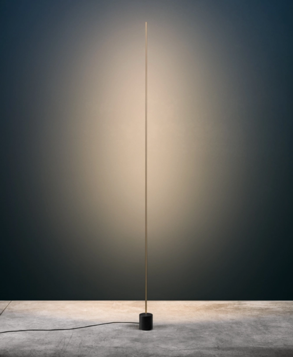 Minimalist floor lamp with a slender gold rod on a black cylindrical base, casting warm vertical light against a dark wall.