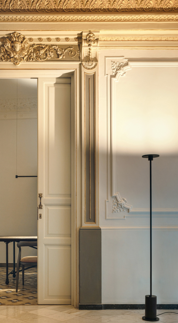Minimalist black floor lamp with a cylindrical base and slim circular head, casting indirect light toward the ceiling in a classic interior adorned with gilded moldings.