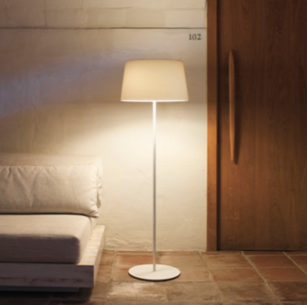 Simple and elegant floor lamp with a slim white stand and beige fabric cylindrical shade, casting a soft glow beside a sofa in a cozy interior.