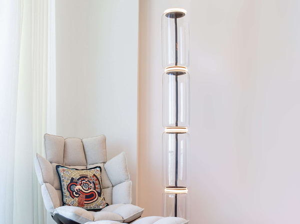 Designer floor lamp made of four stacked transparent glass cylinders connected by LED light rings, set on a wide base, illuminating a reading corner with a beige tufted armchair.