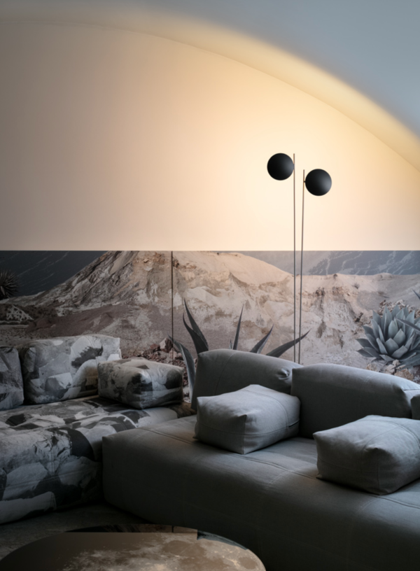 Modern floor lamp with two round black shades, casting warm light onto a vaulted wall, set against a desert landscape mural, with a contemporary grey sofa in the foreground.