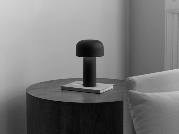 Modern table lamp in matte black metal with a sleek mushroom-shaped design, placed on a round wooden table beside a light-colored sofa.