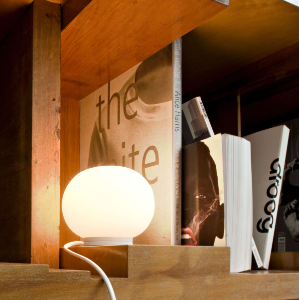 Compact table lamp with a white opaline glass spherical diffuser, emitting a soft glow, placed on a wooden shelf between books.