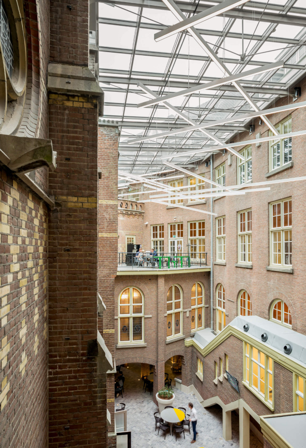 Cour intérieure d’un bâtiment en briques avec toit en verre et suspensions lumineuses linéaires blanches disposées de manière géométrique.