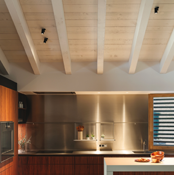 Cuisine moderne avec plafond à poutres apparentes blanches, équipée de projecteurs noirs encastrés en surface, offrant un éclairage ciblé sur le plan de travail et les éléments en inox.