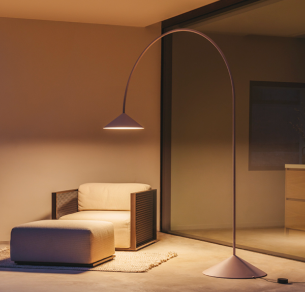 Minimalist arched floor lamp with a conical shade casting warm light, placed next to a beige armchair and ottoman in a contemporary indoor-outdoor space.