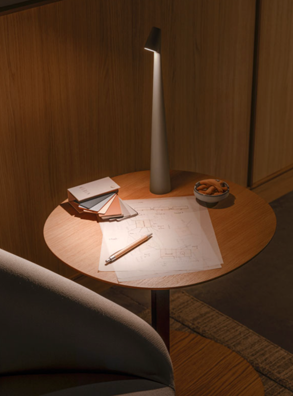 Sleek conical grey metal table lamp with a directional light beam, placed on a round wooden table alongside color swatches and architectural plans.
