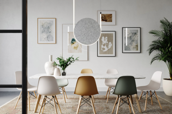 Beige felt circular acoustic pendant hanging above a modern dining table with colorful chairs, in an interior decorated with framed artworks and plants.